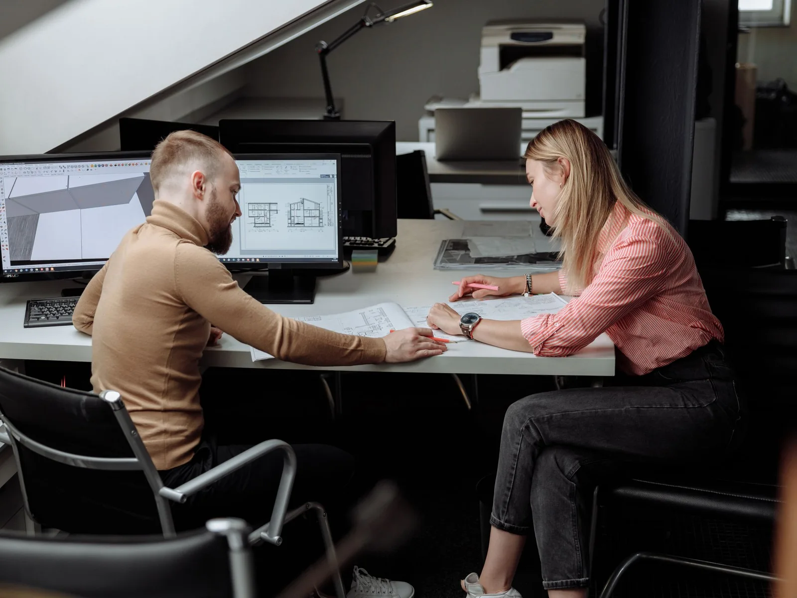 Two professionals reviewing technical drawings at a workstation with computer-aided design screens.