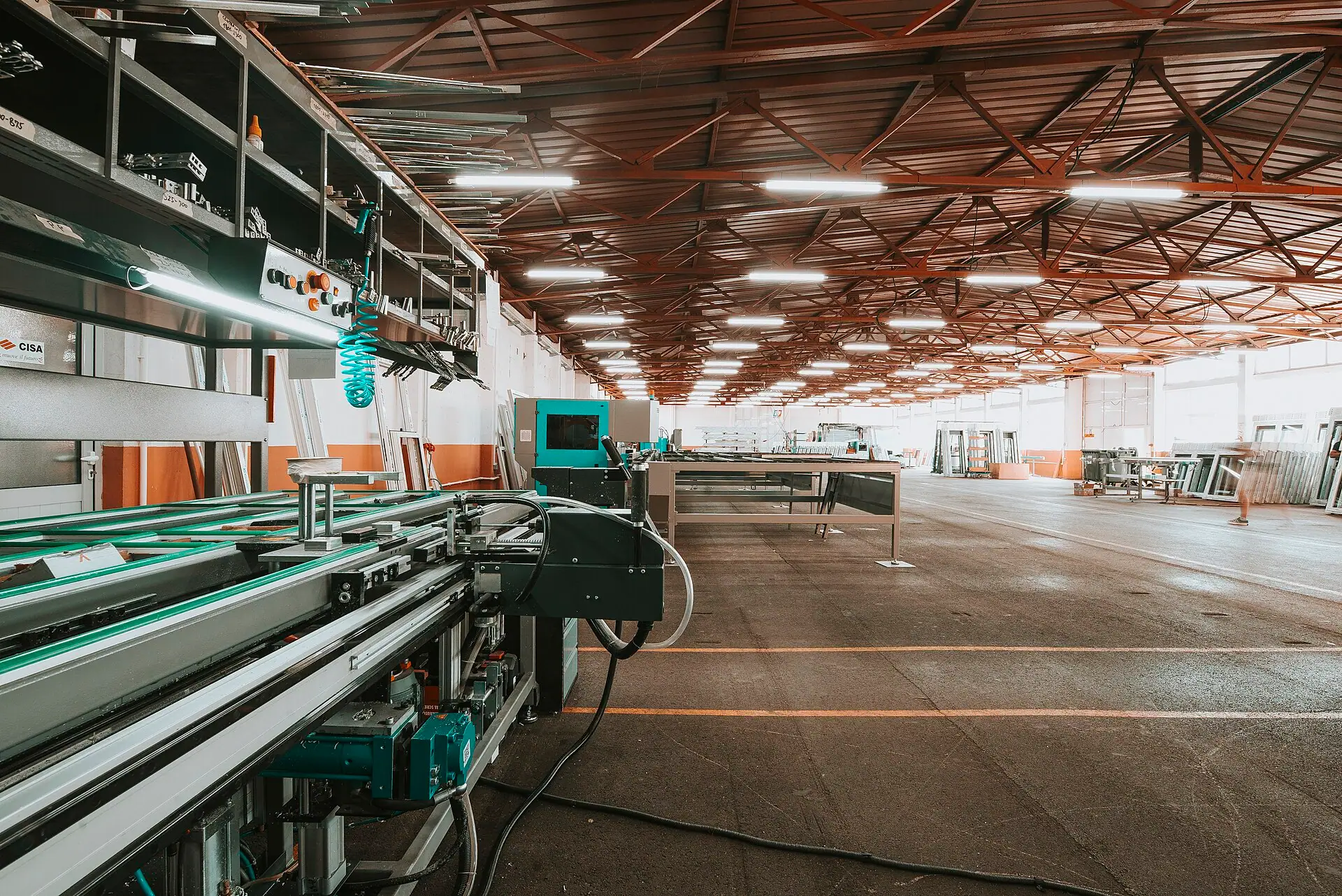 Industrial production floor with processing machinery, metal profiles and overhead services.